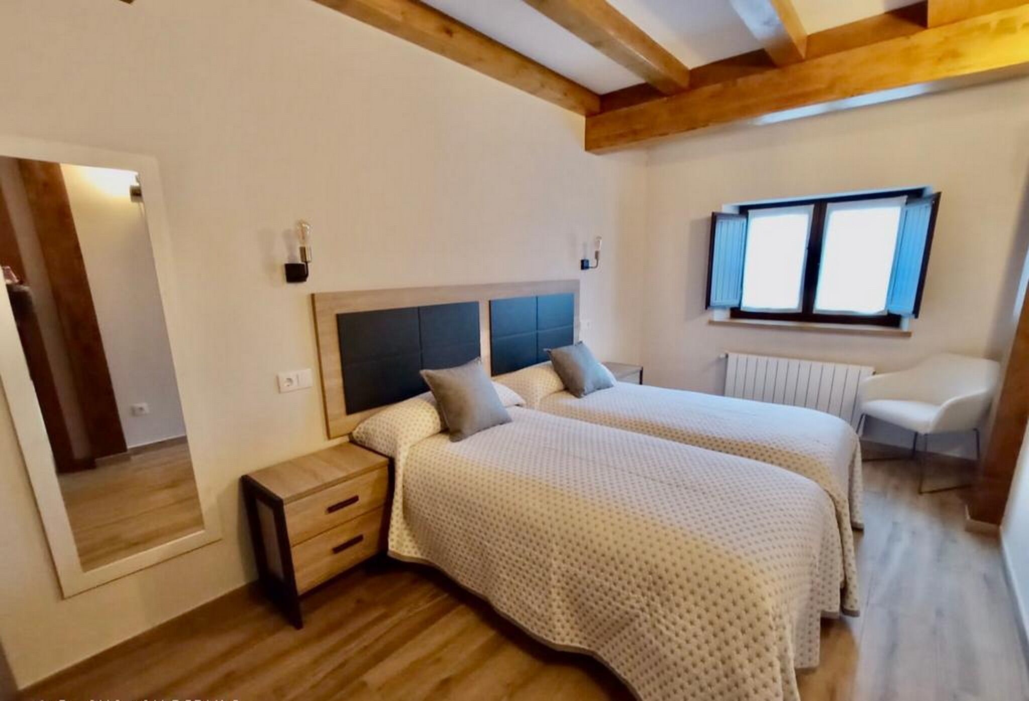 A bedroom with two beds, wooden flooring, a mirror, bedside tables, and a window with blinds.