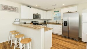 A kitchen with white cabinets, a stainless steel refrigerator, a microwave, and a granite countertop island with bar stools. - Annie by the Sea - Oceanfront, Sunrise & Sunset Decks! (Freeport)