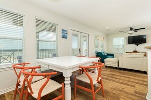 A modern living room with a white dining table, orange chairs, a teal sofa, and a flat-screen TV.