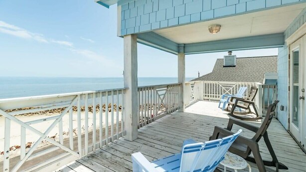 A wooden deck with a railing overlooking a beach, a covered seating area with chairs, and a building with a light blue exterior. - Annie by the Sea - Oceanfront, Sunrise & Sunset Decks! (Freeport)