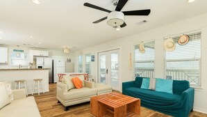 A modern living room with a teal sofa, a wooden coffee table, a white sofa, and a kitchen area in the background. - Annie by the Sea - Oceanfront, Sunrise & Sunset Decks! (Freeport)