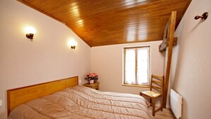 A bedroom with a wooden ceiling, a bed with a beige duvet, a nightstand with a flower vase, a chair, and a window with curtains. - CORINNE - 6 people (ST CREPIN ET CARLUCET)