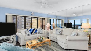A modern living room with two sofas, a coffee table, and a TV. Large windows offer a view of the cityscape. - Rainbow - Ocean Front 1 Bedroom Condo, Steps to the Beach! (Ocean City)