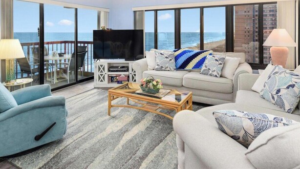 A living room with a sofa, armchair, and coffee table, a television, and a balcony with a view of the ocean. - Rainbow - Ocean Front 1 Bedroom Condo, Steps to the Beach! (Ocean City)
