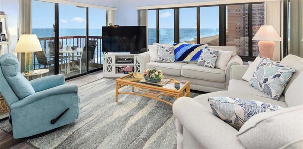 Una sala de estar con sofá, sillón y mesa de centro, televisión y balcón con vistas al océano.