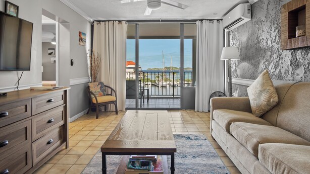 A living room with a wooden coffee table, a beige sofa, a TV on a wooden cabinet, and a view of the sea through a sliding glass door. - Sapphire Dominica-Gorgeous View Walk to beach (St Thomas)