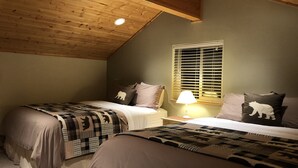 Quarto com duas camas, janela com persianas, abajur em uma mesa de cabeceira e teto de madeira.
