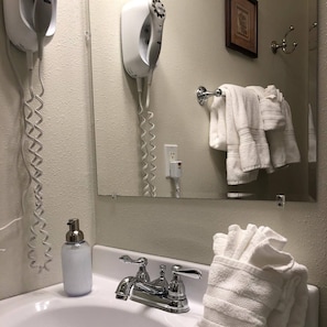 Une salle de bain avec un lavabo blanc, deux robinets, un flacon de savon, un sèche-cheveux et un porte-serviettes avec des serviettes.