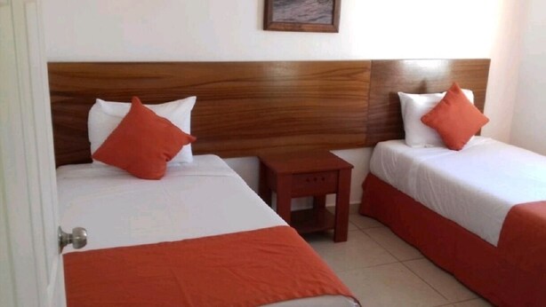 A hotel room with two beds, each with white bedding and orange accents. There is a wooden headboard, a small wooden nightstand, and a framed picture on the wall. - COMFORT DEPARTMENT (Puerto Vallarta)