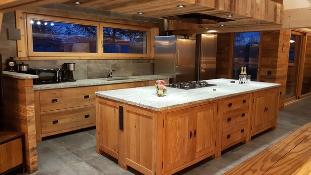 A modern kitchen with wooden cabinetry, a large island, stainless steel appliances, and a view of the outdoors through the windows. - Chalet Pure Paradise 5* (La plagne tarentaise)