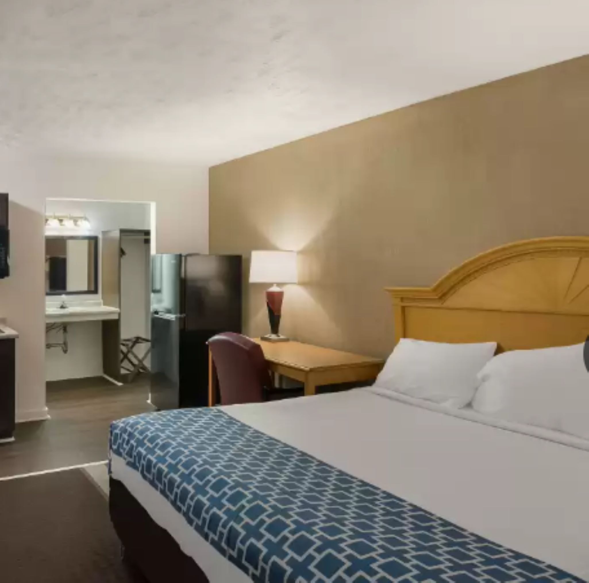 A hotel room with a bed, a desk, a chair, a lamp, a refrigerator, and a mirror.