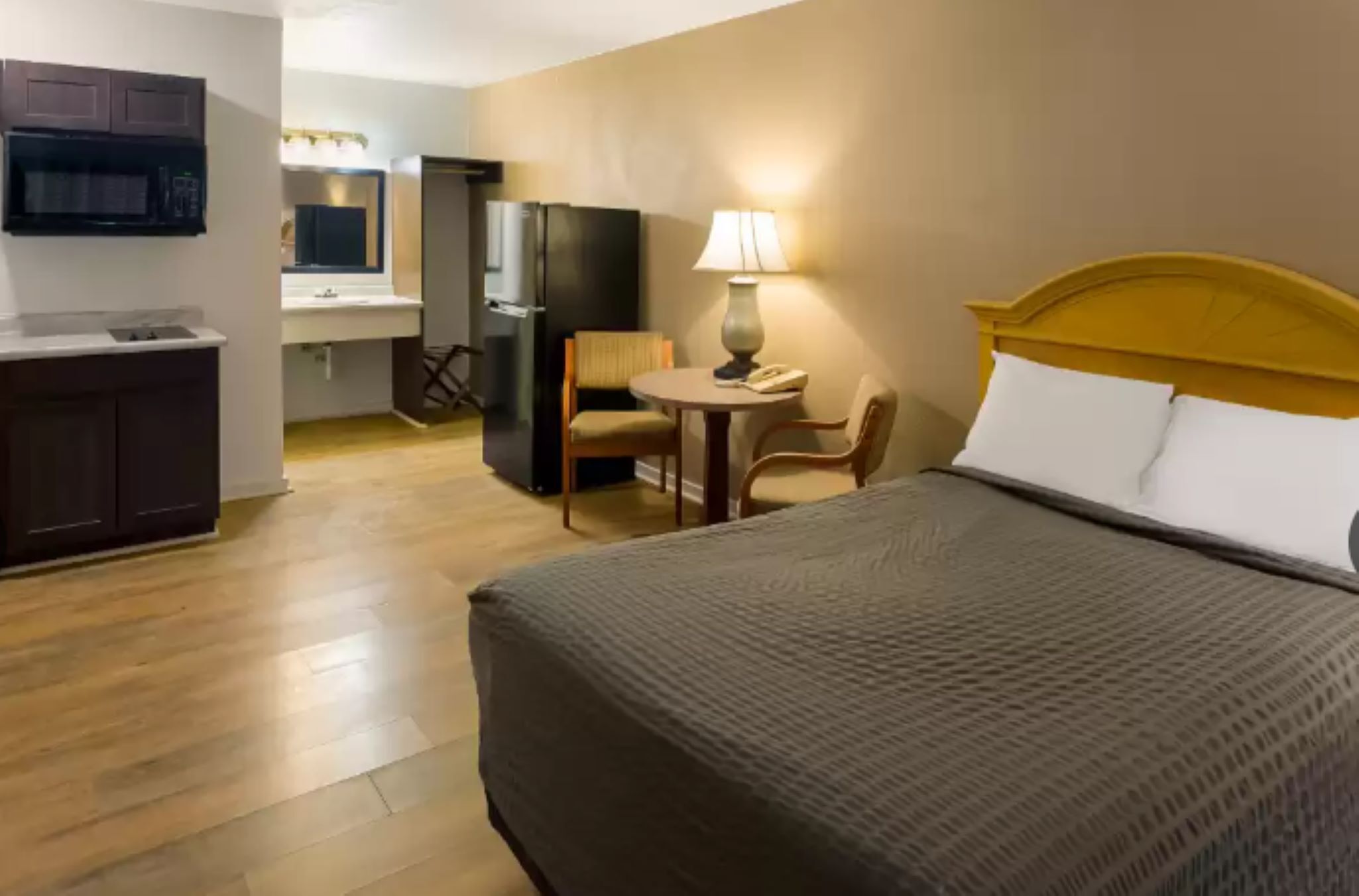 A hotel room with a bed, a desk, a chair, a lamp, and a kitchenette.