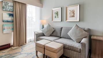 A modern living room with a grey sofa, two matching ottomans, and a large window with beige curtains. There are three framed artworks on the wall and a lamp on a side table.