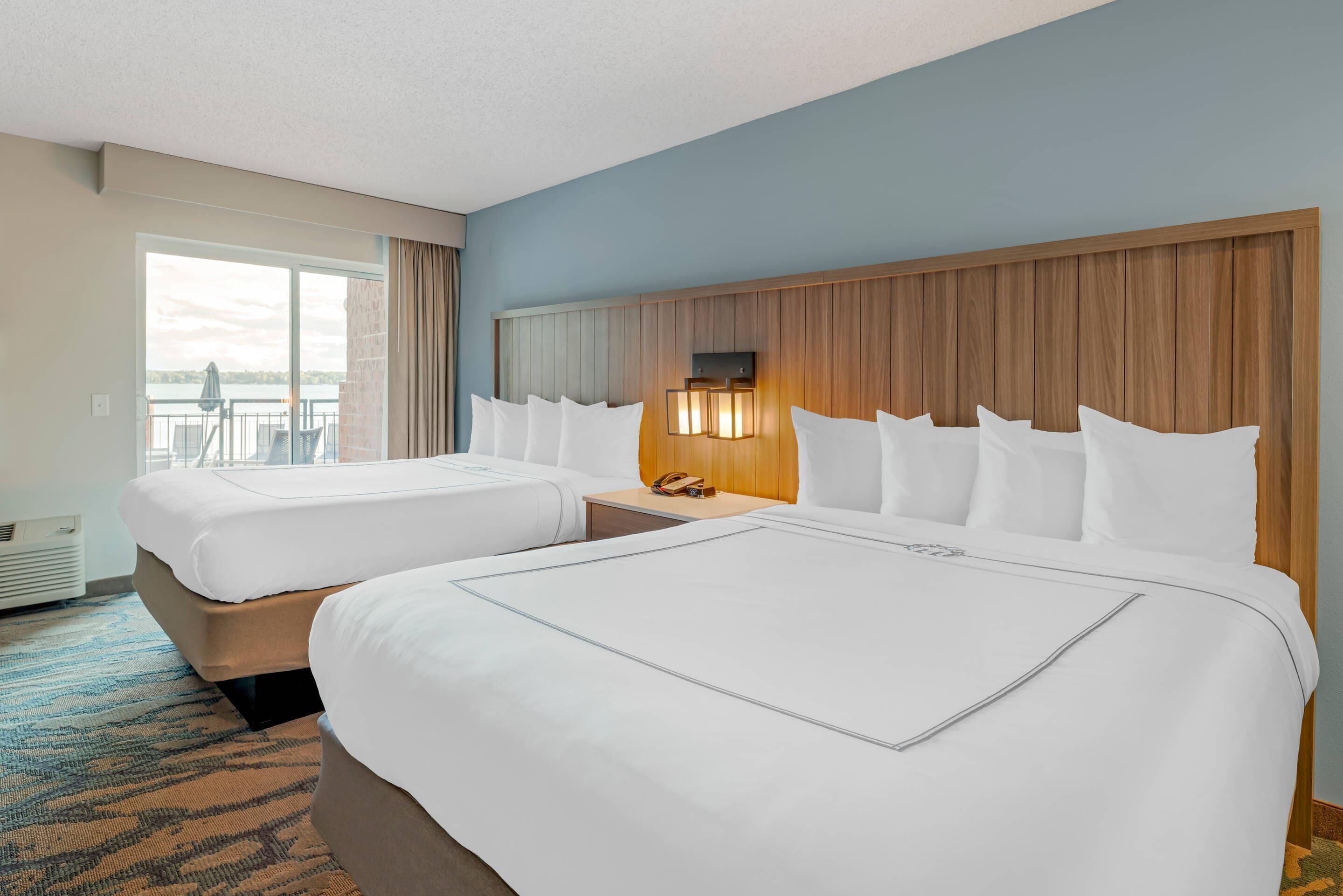 A hotel room with two beds, a wooden headboard, a nightstand with a lamp, and a large window offering a view of the outdoors.
