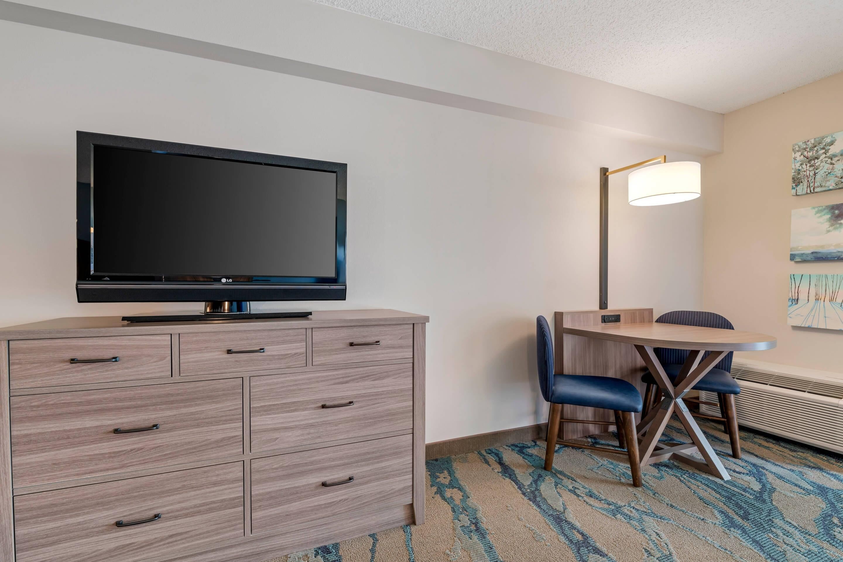 A hotel room with a flat-screen TV on a wooden dresser, a desk with two chairs, a floor lamp, and a large window with a view of trees.