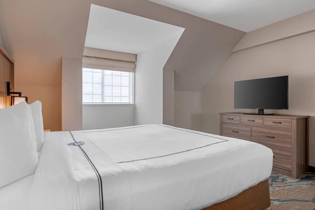 A hotel room with a large bed, a TV on a wooden dresser, a window with a view, and a skylight.