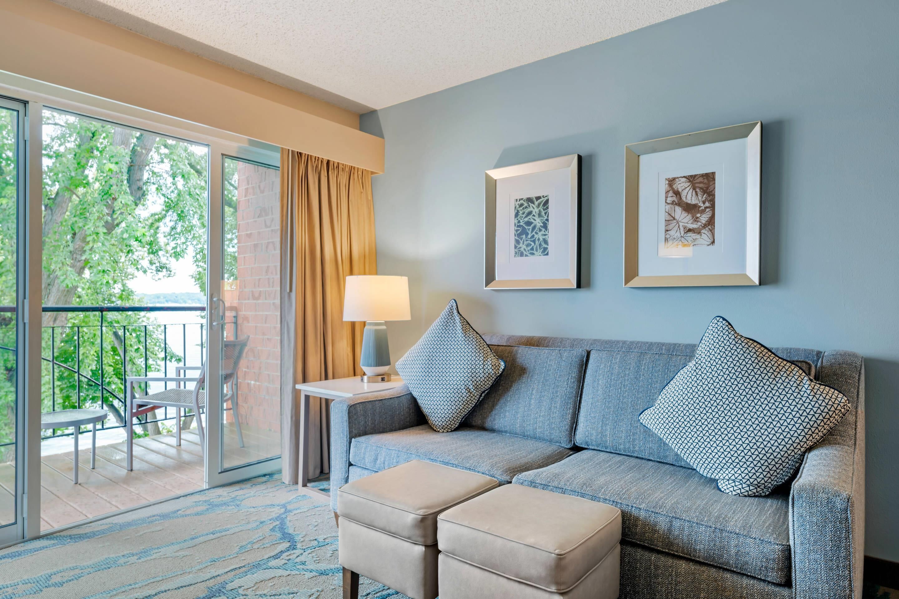 A hotel room with a sofa, ottoman, and a balcony with outdoor furniture.