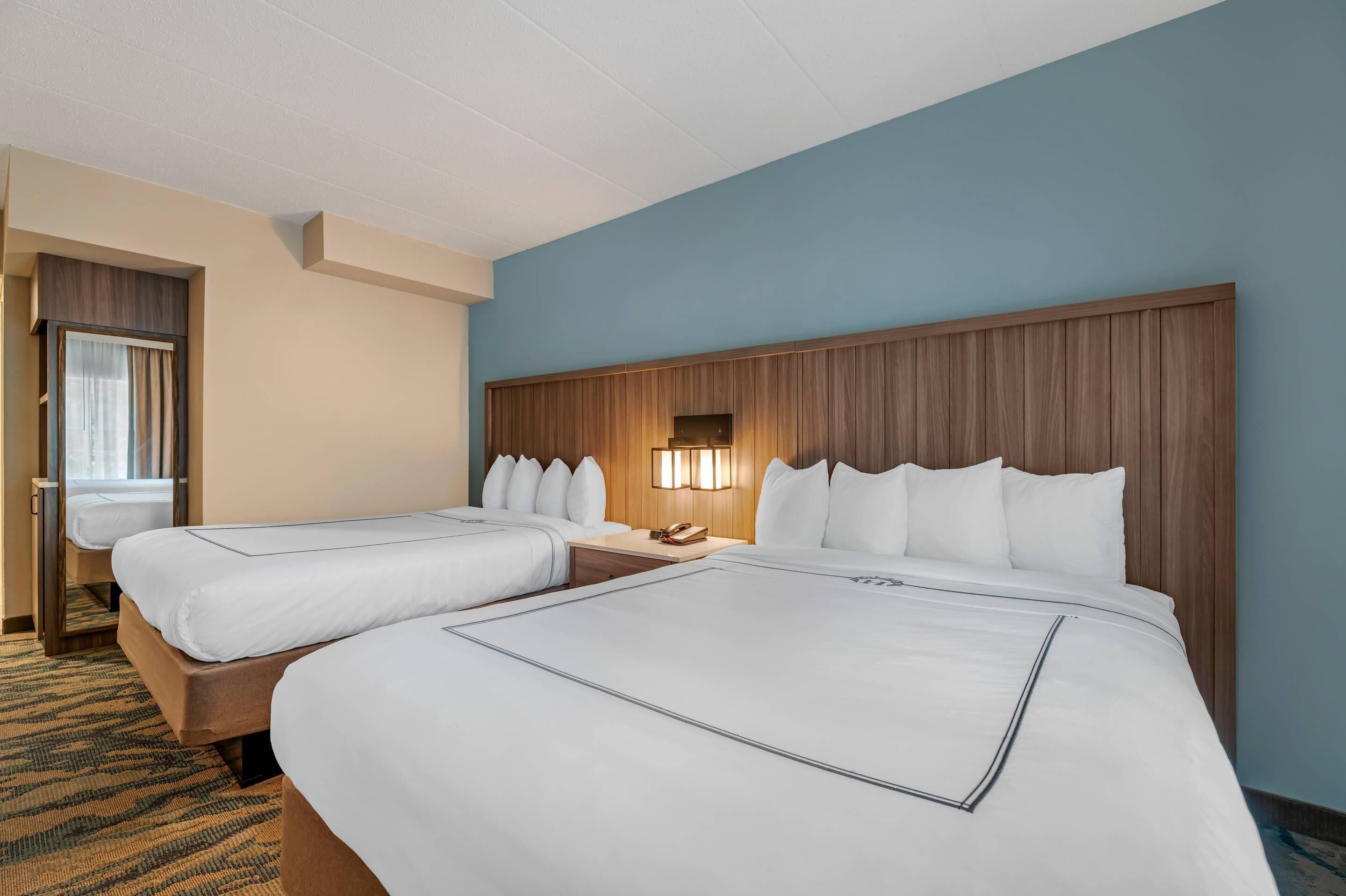 Habitación de hotel con dos camas, cabecero de madera, mesita de noche con lámpara y ventana con cortinas.