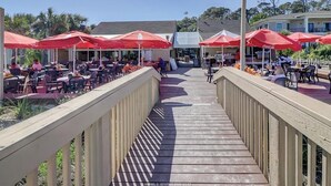 Een houten terras met buitenmeubilair en rode parasols, met uitzicht op een zwembad en een gebouw met een wit dak.