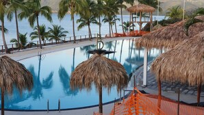 A pool with palm trees and thatched umbrellas, surrounded by a fence and a view of the sea.