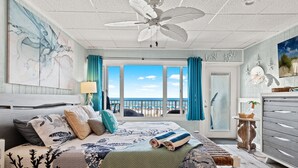 Un dormitorio con una cama grande, ventilador de techo, vista a la playa y puerta que da a un balcón.