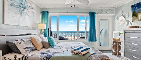 A bedroom with a large bed, a ceiling fan, a view of the beach, and a door leading to a balcony.