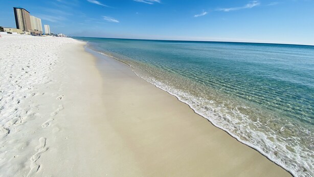 Una playa con agua cristalina, una orilla arenosa y edificios al fondo.