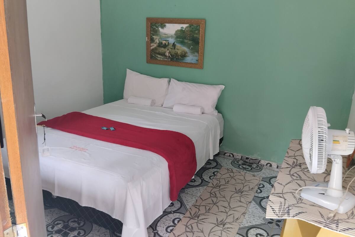 Een klein, netjes opgemaakt bed met witte lakens en een rode deken, een aan de muur hangend landschapsbeeld, en een witte ventilator op een tafel met dessin.