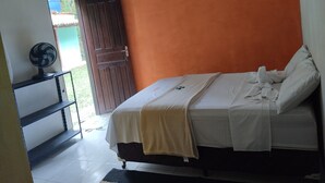 Een slaapkamer met een oranje muur, een bed met witte lakens, een ventilator op een plank en een deur die naar buiten leidt.
