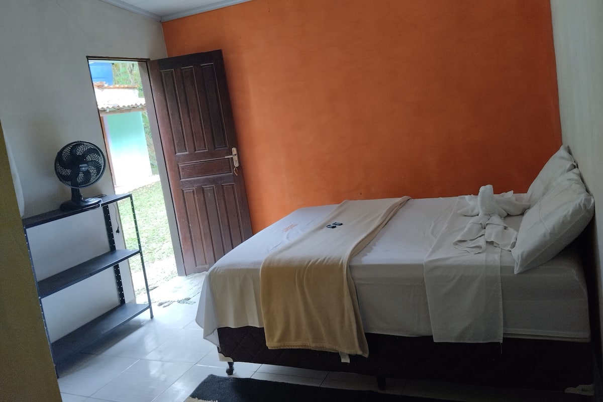 Een slaapkamer met een oranje muur, een bed met witte lakens, een ventilator op een plank en een deur die naar buiten leidt.