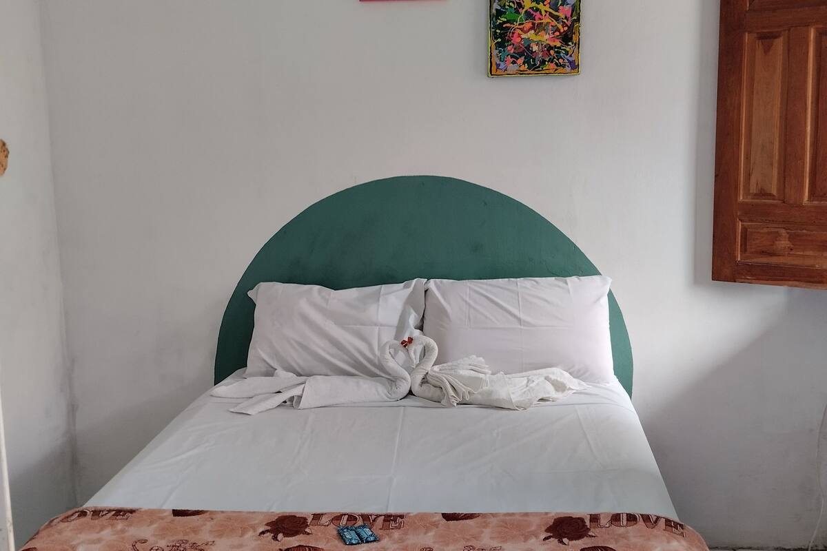 Een net opgemaakt bed met witte lakens en een tealkleurig hoofdbord, een witte handdoek en een bruine deken met de tekst 'LOVE' geborduurd. Boven het bed hangen drie kleurrijke schilderijen.