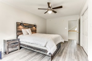 A bedroom with a wooden headboard, a bed with white bedding, a nightstand, a ceiling fan, and a door leading to another room.