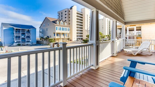 A wooden deck with a railing, a lounge chair, and a table. In the background, there are multi-story buildings with balconies.