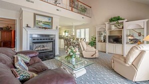 A spacious living room with a fireplace, leather sofas, a glass coffee table, and a high ceiling with a balcony.