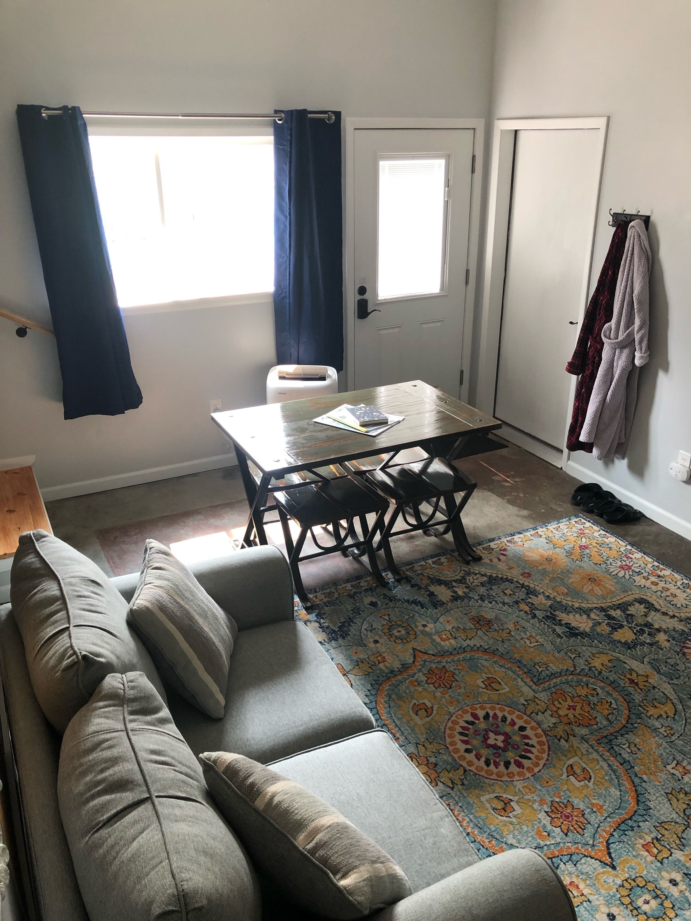 A living room with a sofa, a table with chairs, a patterned rug, and a door with curtains.