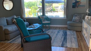 A living room with a grey sofa, a wicker chair, a blue armchair, a round woven rug, and a view of a pool and buildings through large windows.
