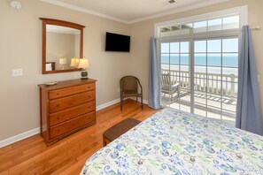 A bedroom with a wooden dresser, a chair, a bed with a floral bedspread, a large mirror, a flat-screen TV, and a balcony with a view of the ocean.