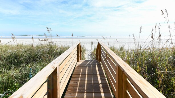 Ein hölzerner Steg führt zu einem Strand mit blauem Himmel.