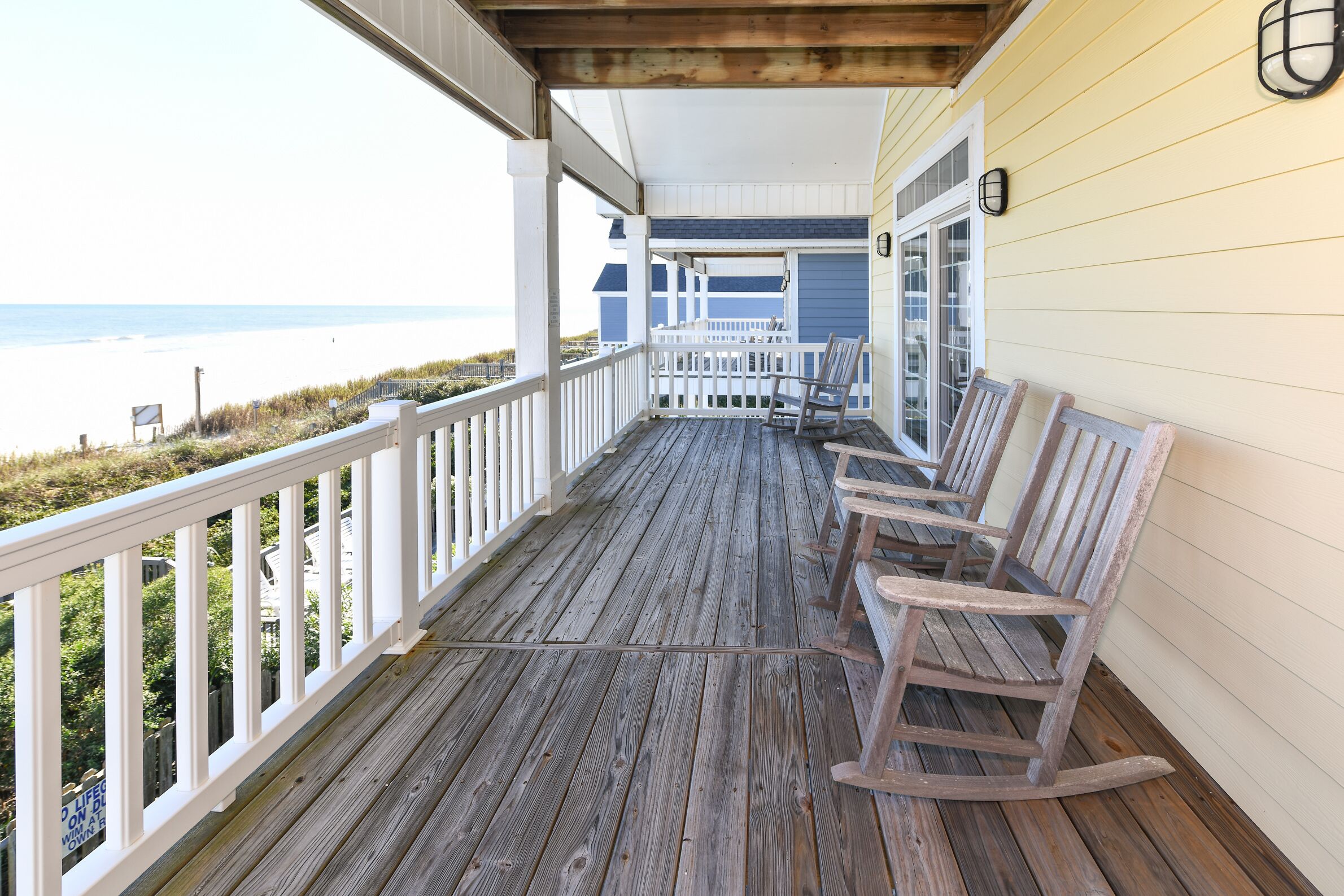 Une terrasse en bois avec des balustrades blanches et des fauteuils à bascule, donnant sur une plage.