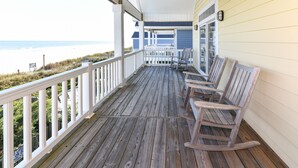 Een houten terras met witte leuningen en schommelstoelen, met uitzicht op een strand.