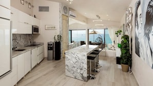 A modern kitchen with marble countertops, a central island with bar stools, a ceiling fan, and a view of the sea through large windows.