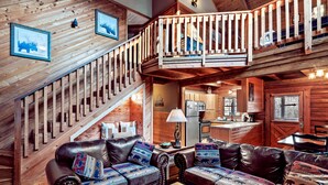 A rustic cabin interior with a wooden staircase, leather sofas with patterned cushions, a wooden dining table, and a kitchen area.