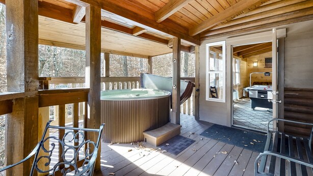 A wooden deck with a hot tub, outdoor seating, and a view of the surrounding forest.