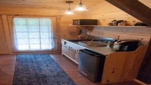 A compact kitchen with wooden walls, a sink, and a dishwasher. There is a window with curtains, a blue rug, and a ceiling light fixture. - Masons Chapel at Springfield 1842 (Halifax)