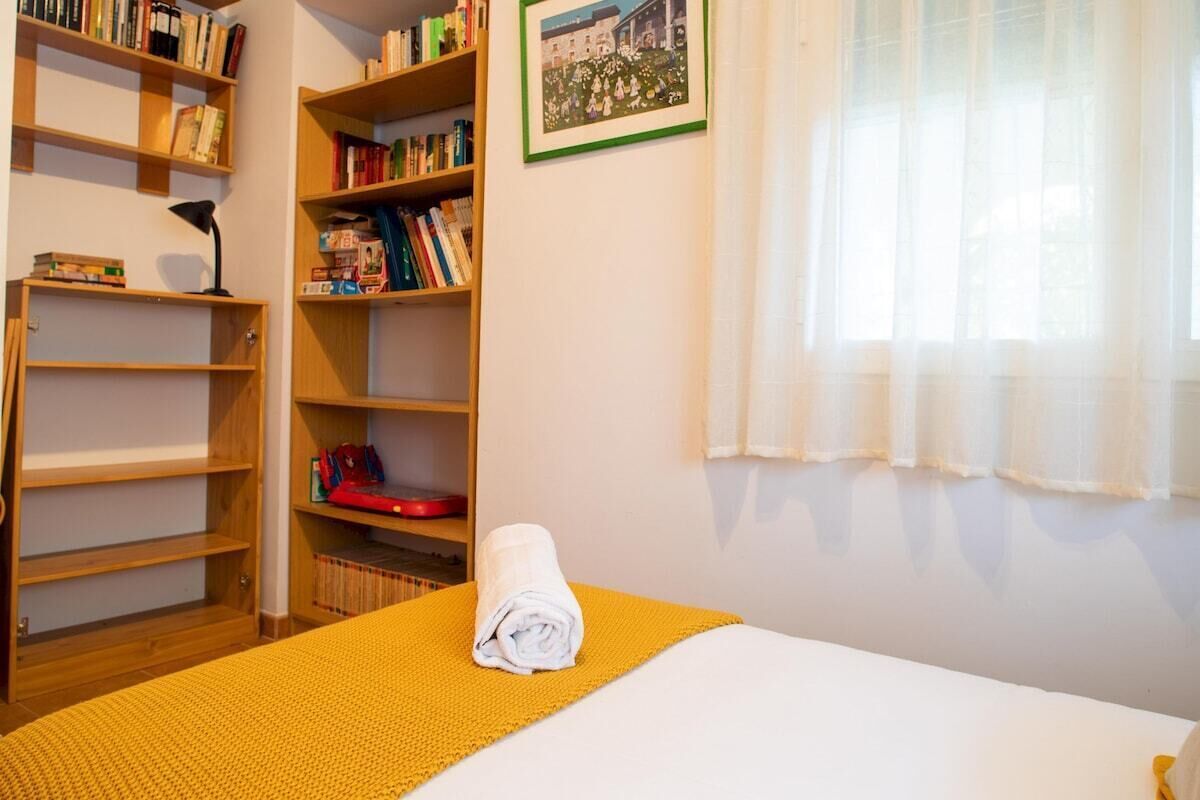 Habitació d'hotel amb un llit, una manta groga, una tovallola blanca arrotllada i prestatges de fusta amb llibres i un objecte vermell.