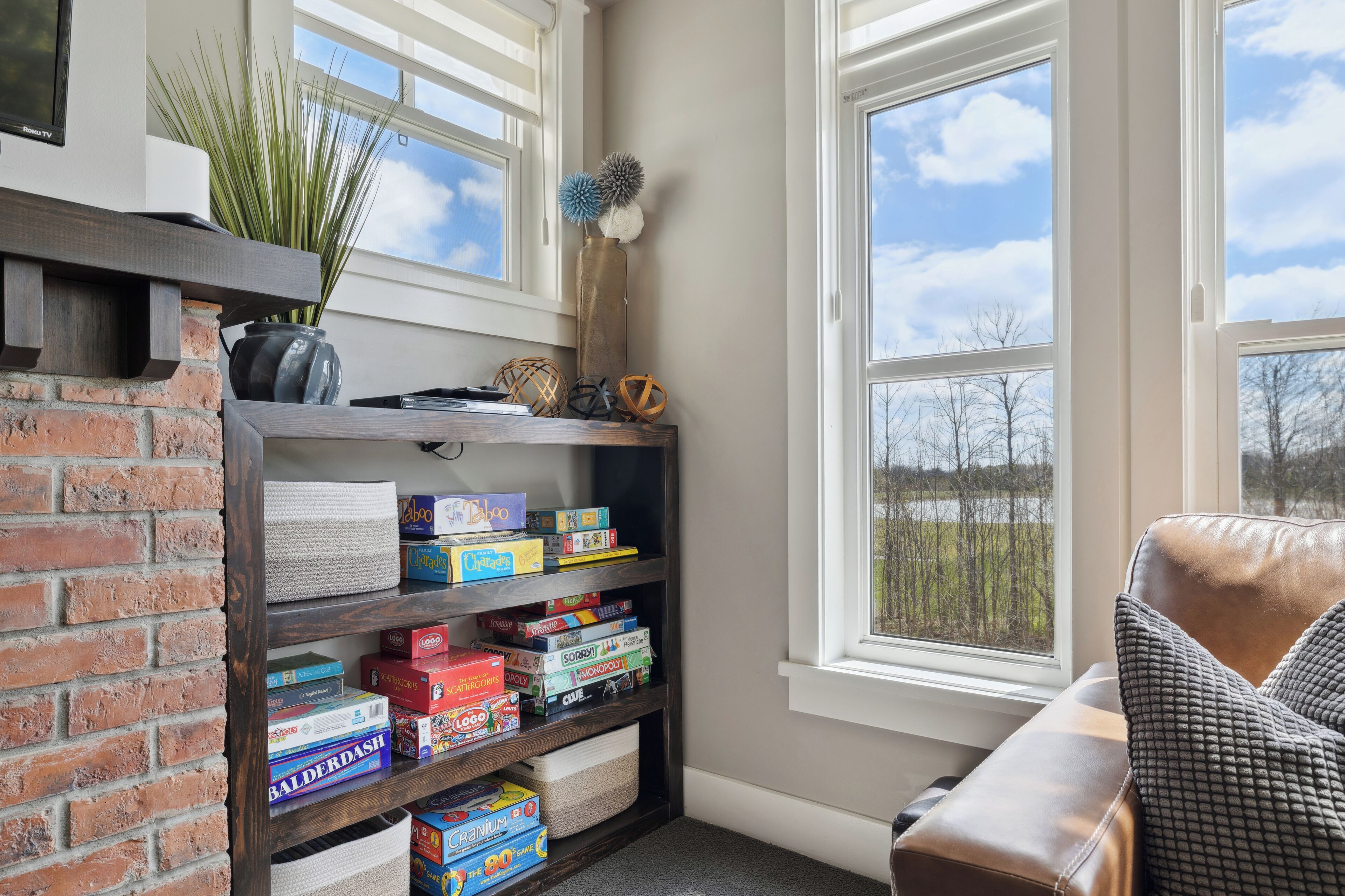 A corner room with a brick fireplace, a bookshelf with board games, a window with a view of trees, and a leather chair.