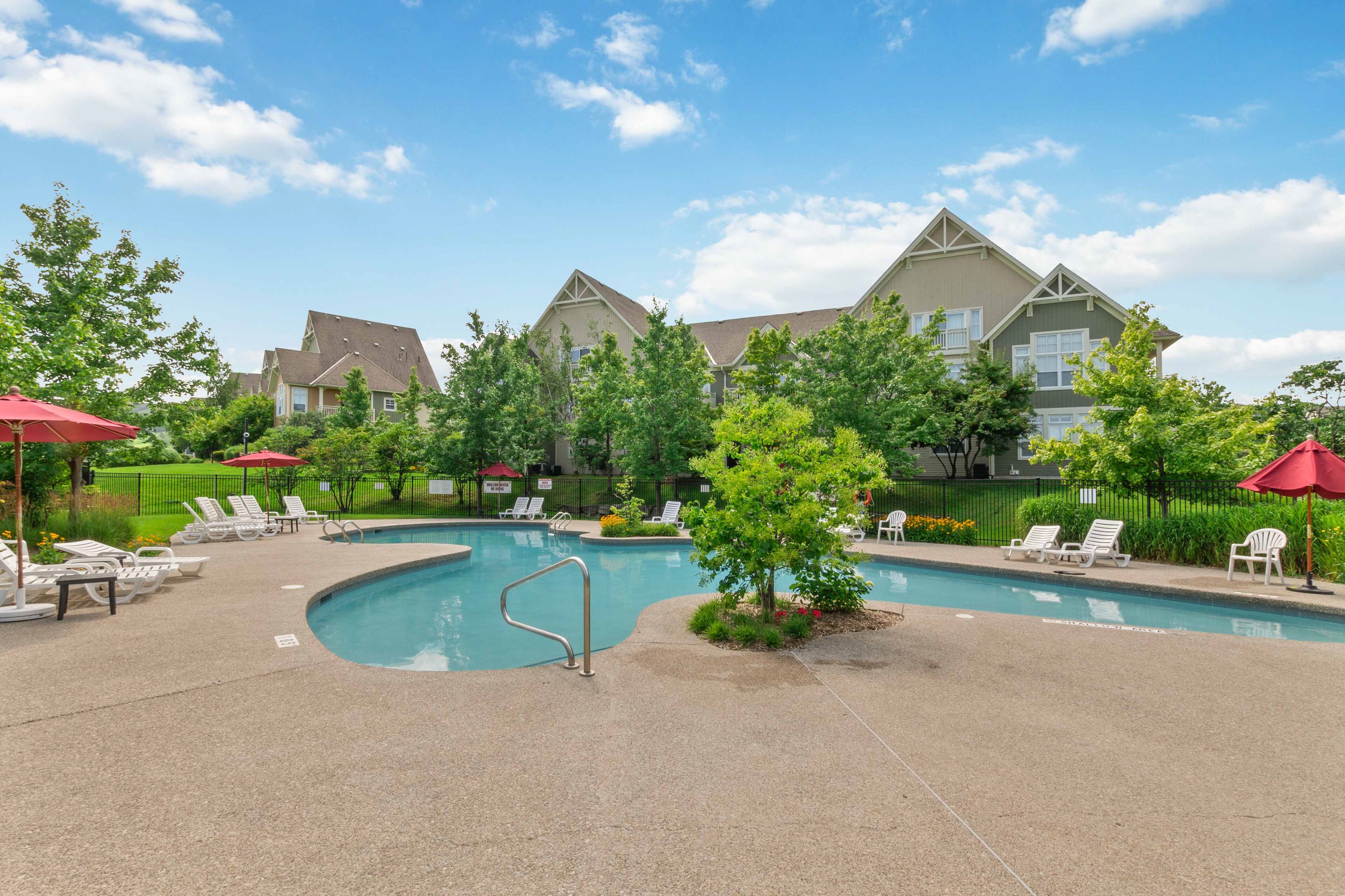 A swimming pool with lounge chairs and umbrellas, surrounded by greenery and a building with a pitched roof.