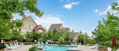 A swimming pool with lounge chairs and umbrellas, surrounded by trees and buildings.
