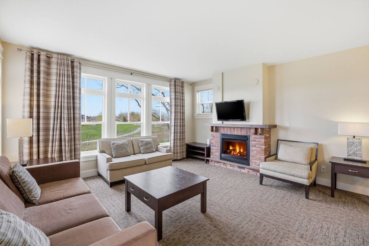 A living room with a sofa, armchair, coffee table, fireplace, TV, and large windows with curtains.