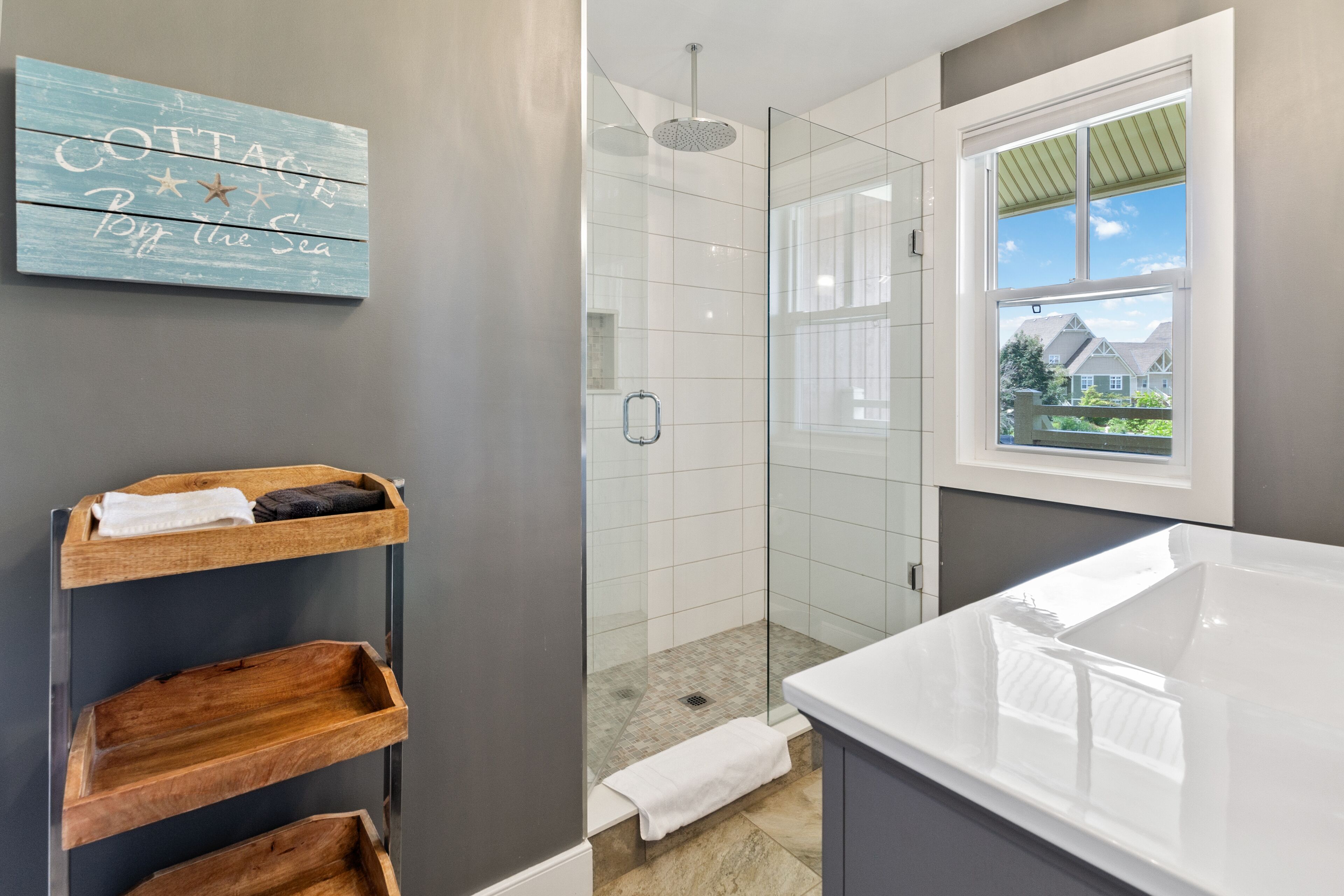 A modern bathroom with a glass shower enclosure, a wooden shelf with towels, and a view of houses through a window.